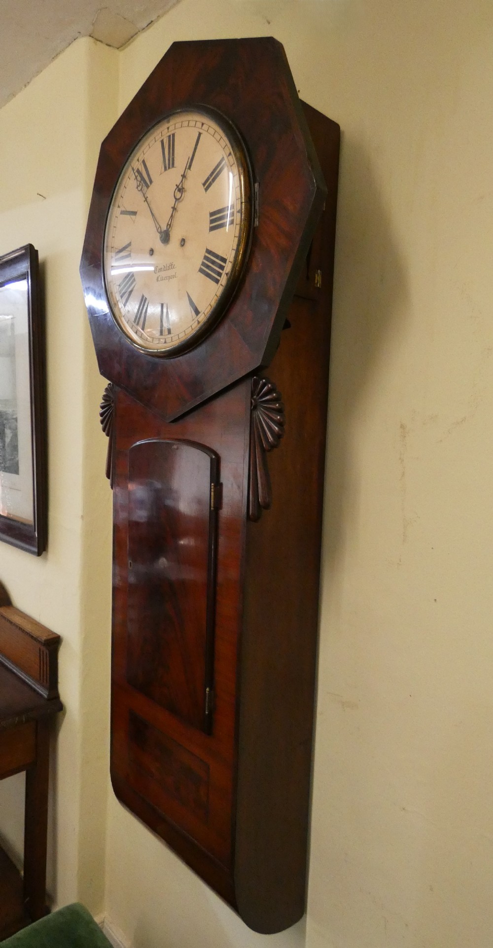 An antique mahogany tavern wall clock featuring an octagonal dial surround, carved side ears, and a long rectangular trunk with a flame mahogany door. View V