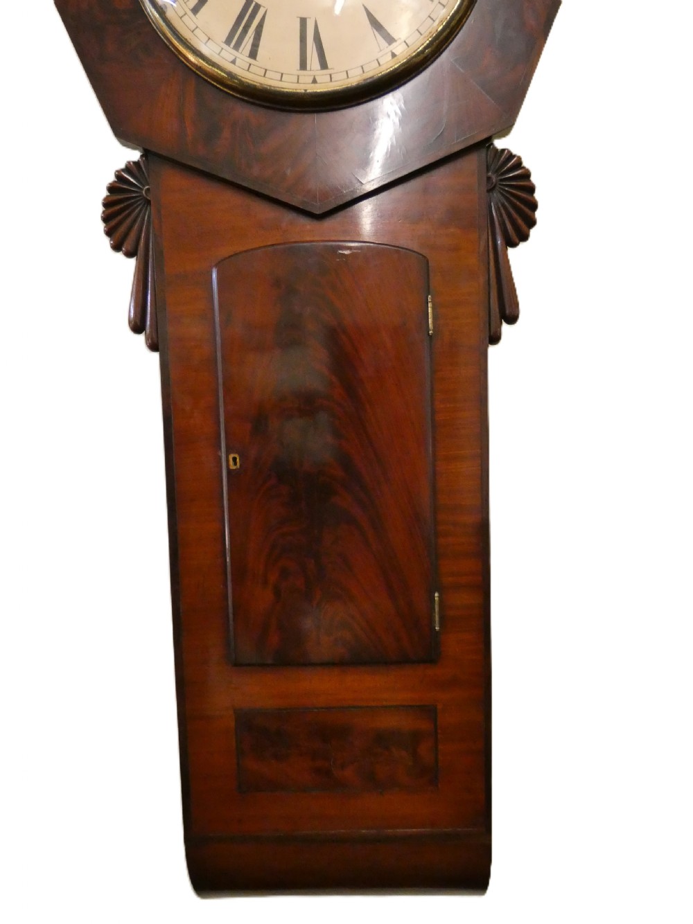An antique mahogany tavern wall clock featuring an octagonal dial surround, carved side ears, and a long rectangular trunk with a flame mahogany door. View VII