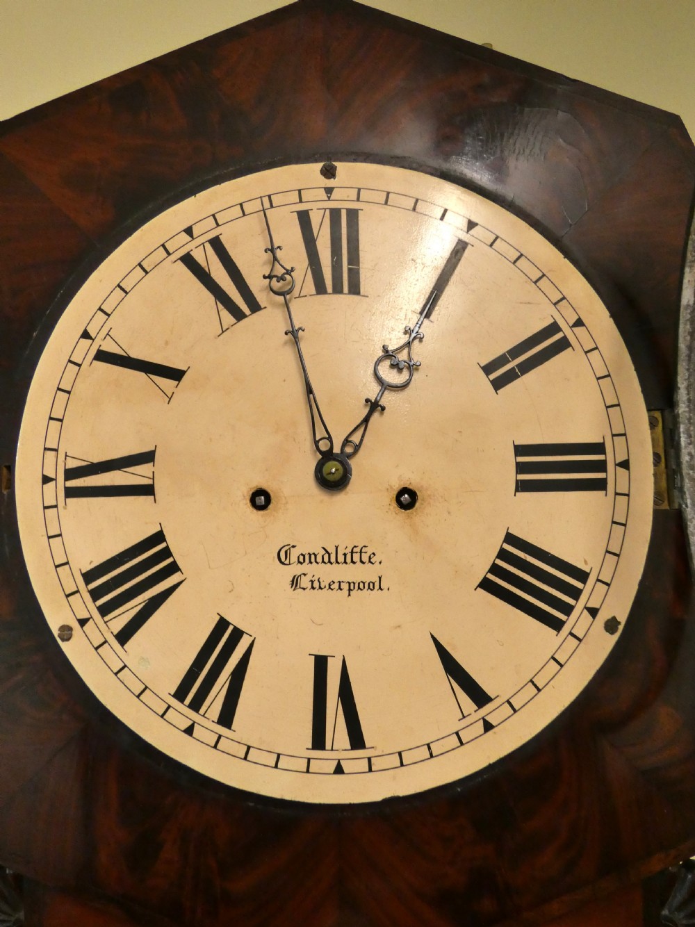 An antique mahogany tavern wall clock featuring an octagonal dial surround, carved side ears, and a long rectangular trunk with a flame mahogany door. View VIII
