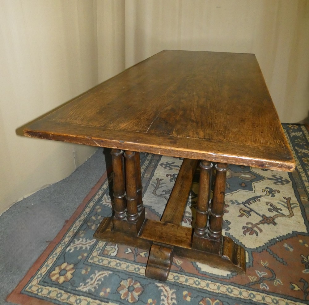 An antique oak refectory table featuring a rectangular top resting on clustered turned column trestle supports joined by a central floor stretcher. View II