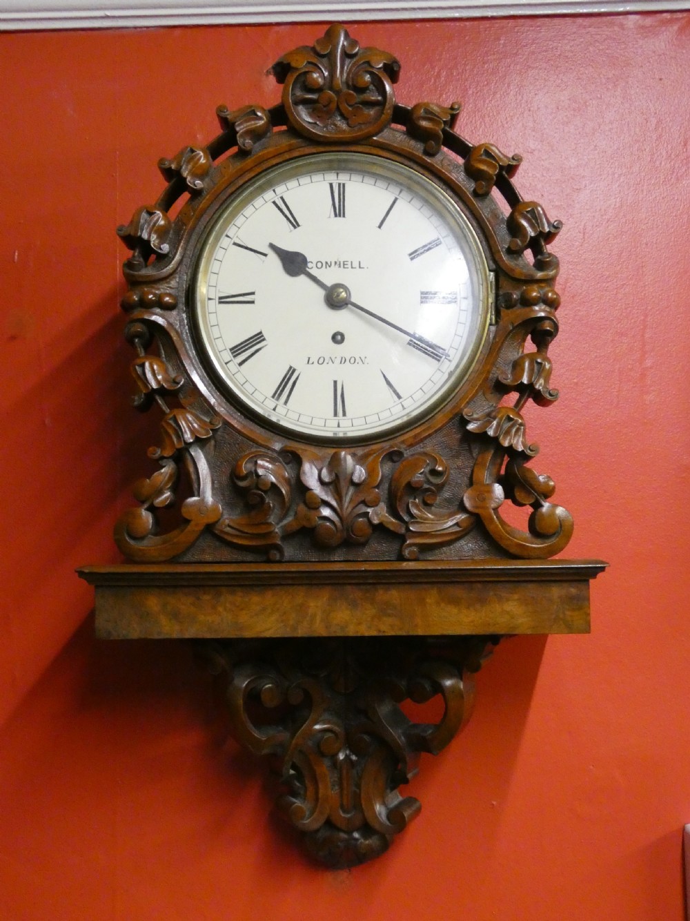 A carved walnut bracket clock featuring scrollwork ornamentation, a wall-mounting bracket, and a white dial displaying Roman numerals with the maker name Connell London.