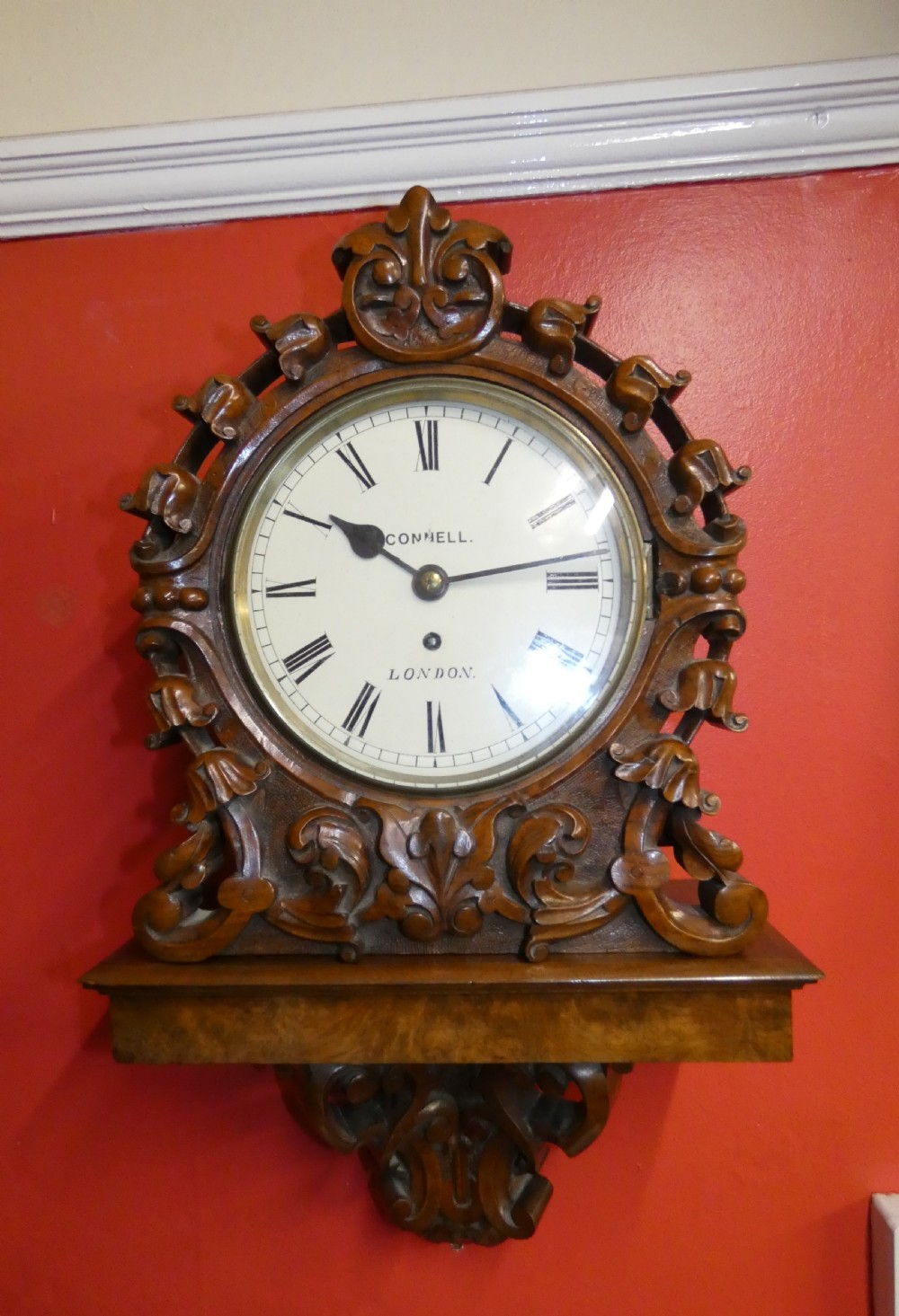 A carved walnut bracket clock featuring scrollwork ornamentation, a wall-mounting bracket, and a white dial displaying Roman numerals with the maker name Connell London. View II