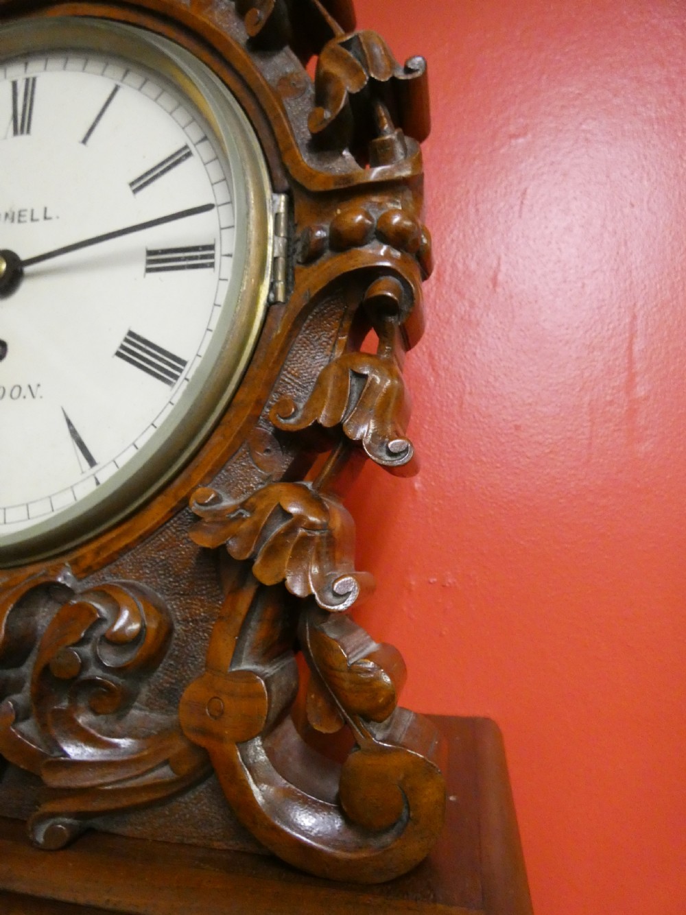 A carved walnut bracket clock featuring scrollwork ornamentation, a wall-mounting bracket, and a white dial displaying Roman numerals with the maker name Connell London. View III