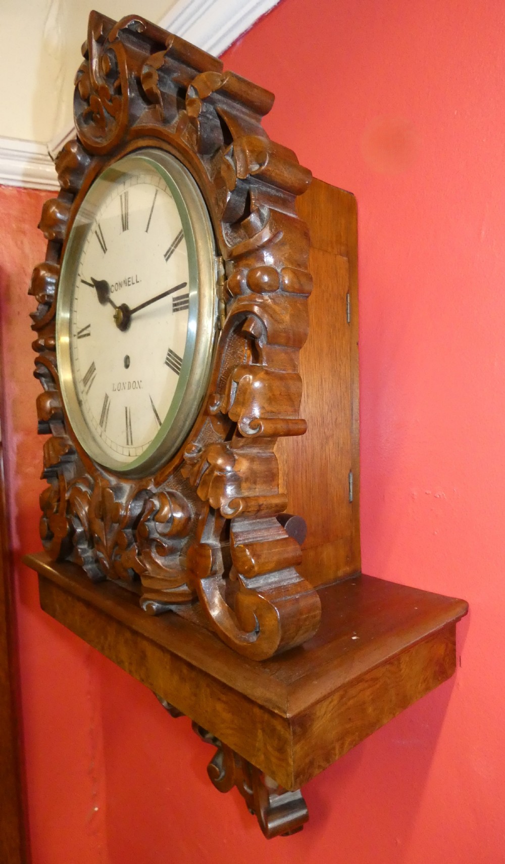 A carved walnut bracket clock featuring scrollwork ornamentation, a wall-mounting bracket, and a white dial displaying Roman numerals with the maker name Connell London. View V