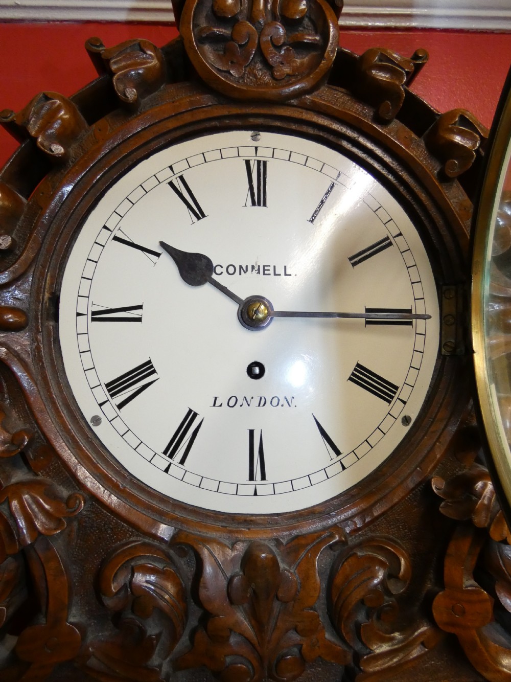 A carved walnut bracket clock featuring scrollwork ornamentation, a wall-mounting bracket, and a white dial displaying Roman numerals with the maker name Connell London. View VI