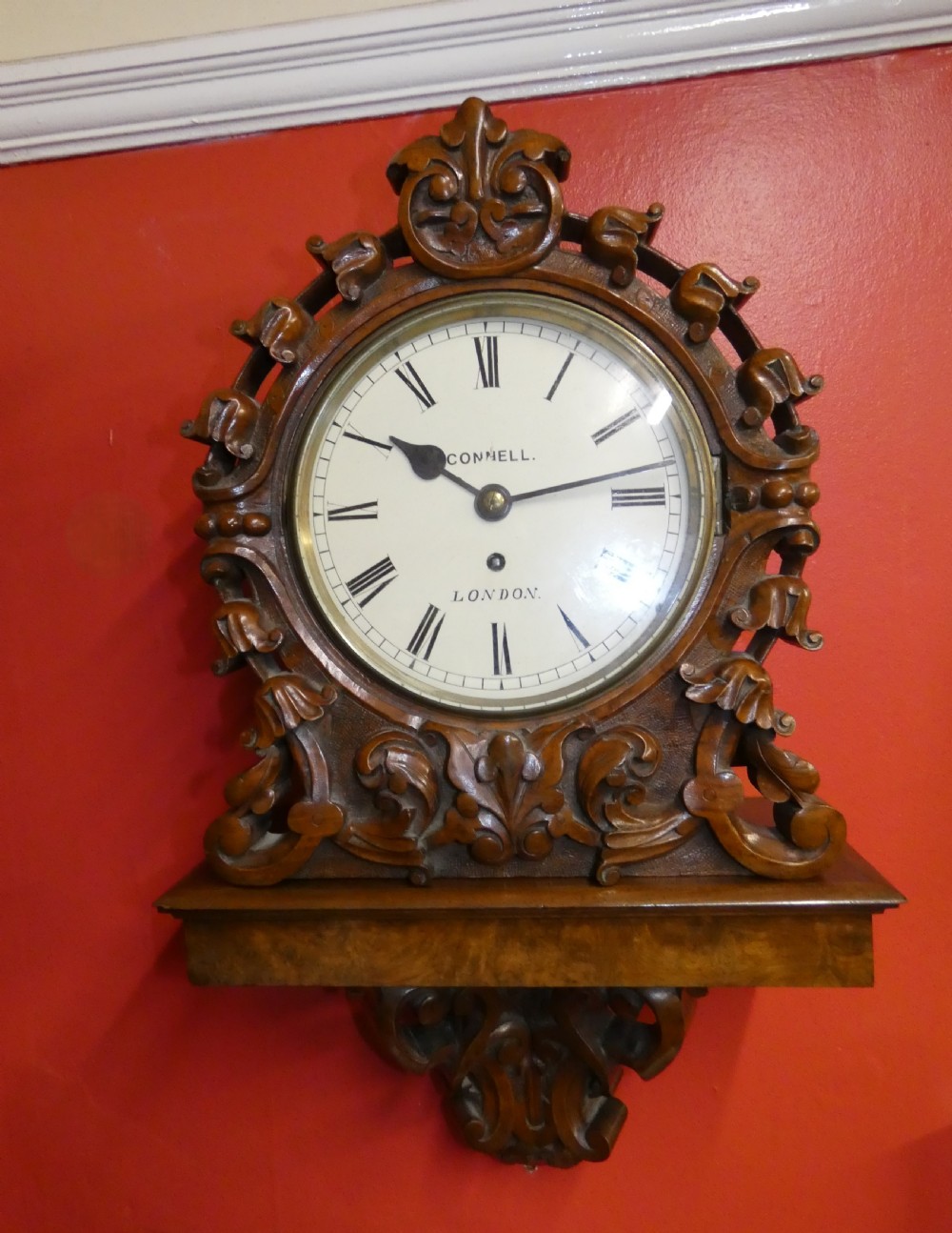 A carved walnut bracket clock featuring scrollwork ornamentation, a wall-mounting bracket, and a white dial displaying Roman numerals with the maker name Connell London. View IX