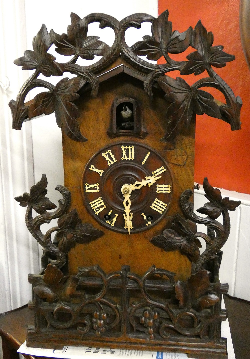 Hand-carved dark wood Black Forest cuckoo clock featuring intricate vine leaf ornamentation around a circular dial with Roman numerals and a cuckoo bird aperture.
