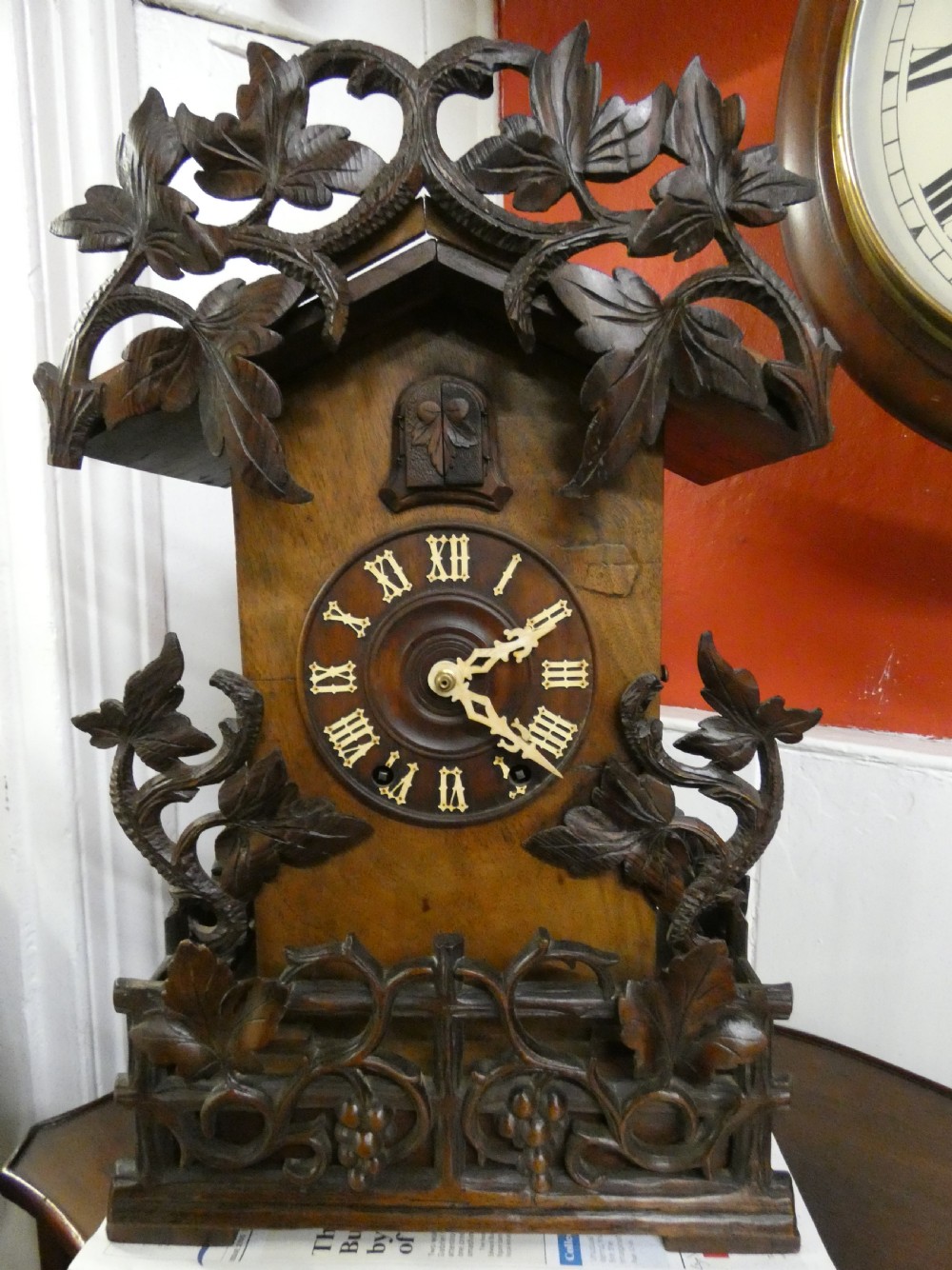 Hand-carved dark wood Black Forest cuckoo clock featuring intricate vine leaf ornamentation around a circular dial with Roman numerals and a cuckoo bird aperture. View II