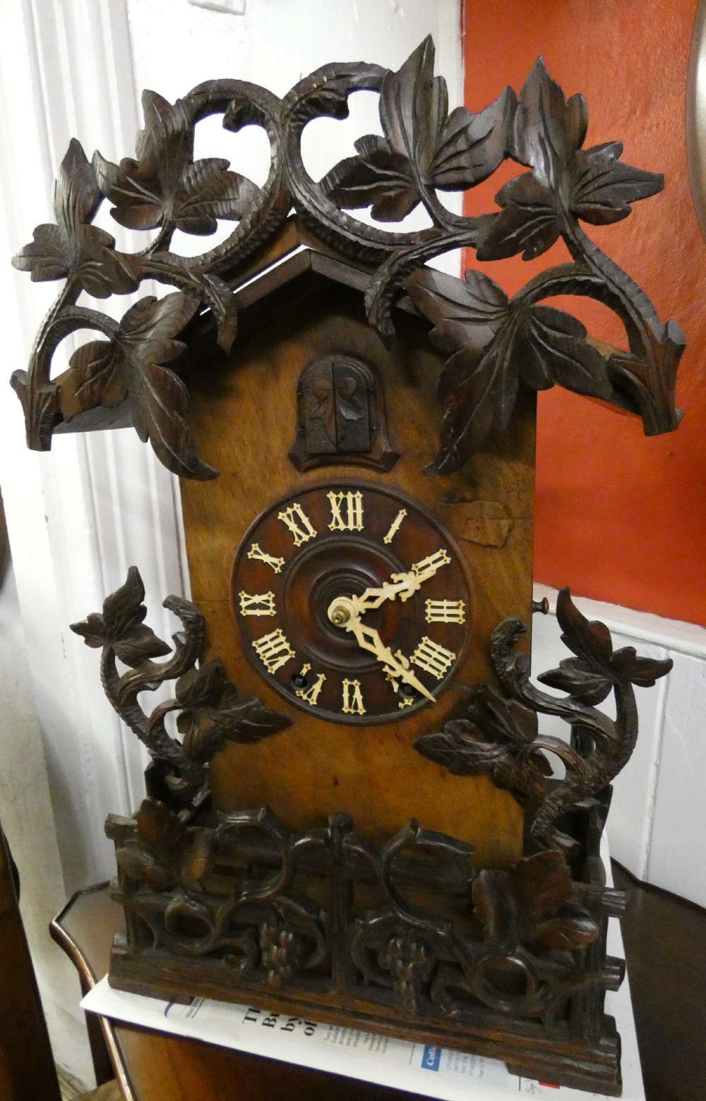 Hand-carved dark wood Black Forest cuckoo clock featuring intricate vine leaf ornamentation around a circular dial with Roman numerals and a cuckoo bird aperture. View III