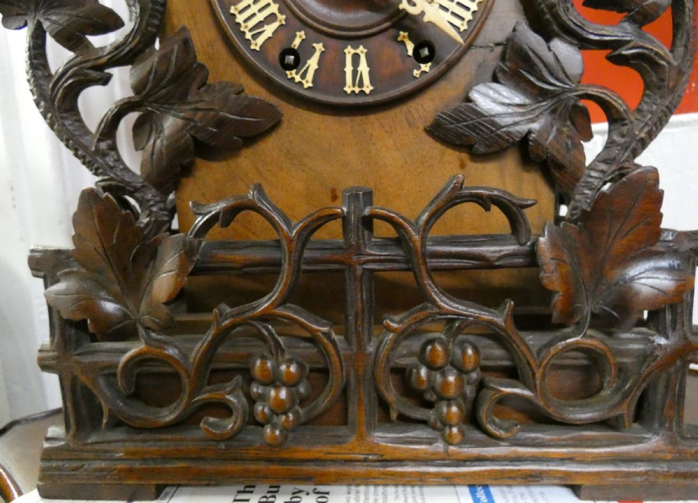Hand-carved dark wood Black Forest cuckoo clock featuring intricate vine leaf ornamentation around a circular dial with Roman numerals and a cuckoo bird aperture. View IV