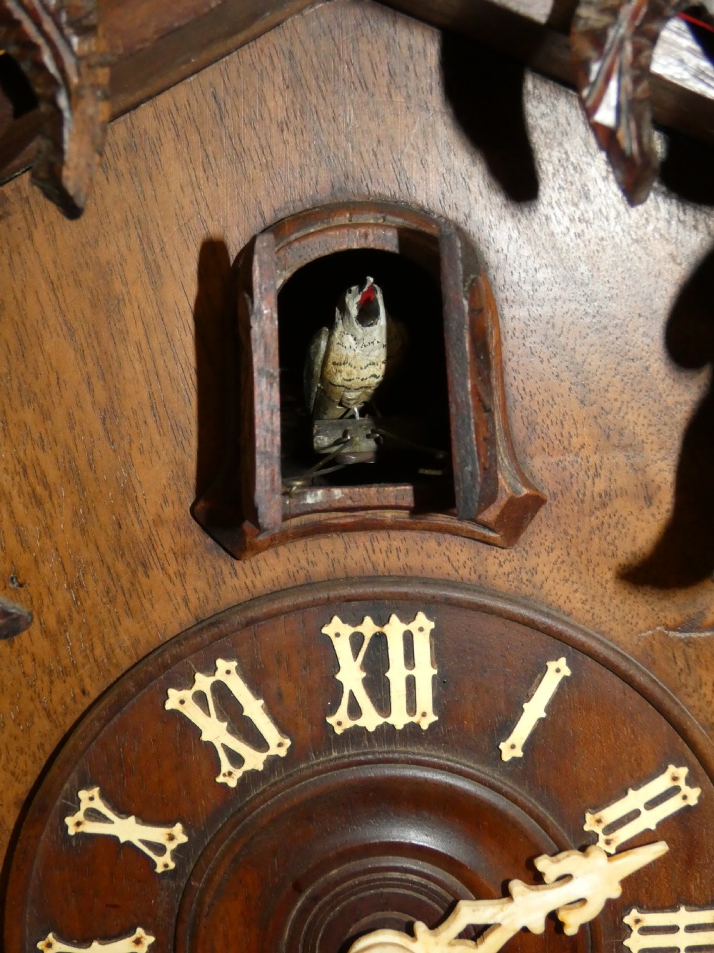 Hand-carved dark wood Black Forest cuckoo clock featuring intricate vine leaf ornamentation around a circular dial with Roman numerals and a cuckoo bird aperture. View IX