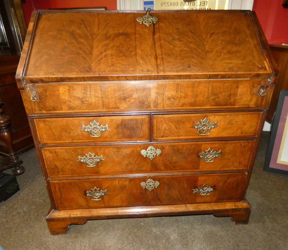 A George II period walnut slant-front bureau featuring figured veneers, brass drop handles, keyhole escutcheons, and graduating drawers on bracket feet.