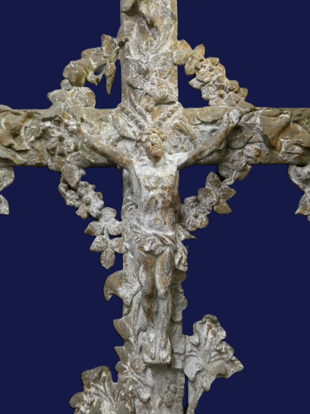 A weathered cast iron grave crucifix featuring detailed foliate work along the vertical beam and the figure of Christ in relief against a distressed grey finish. View VI