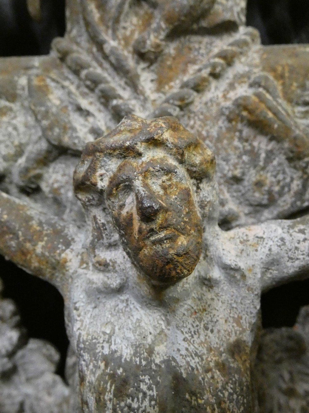 A weathered cast iron grave crucifix featuring detailed foliate work along the vertical beam and the figure of Christ in relief against a distressed grey finish. View VII