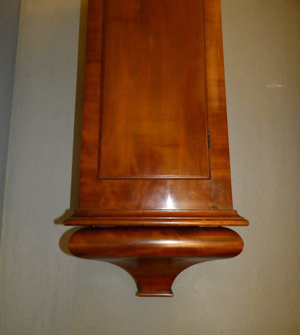 A tall Victorian mahogany regulator wall clock featuring a circular white dial with Roman numerals and a long rectangular wooden trunk. View II