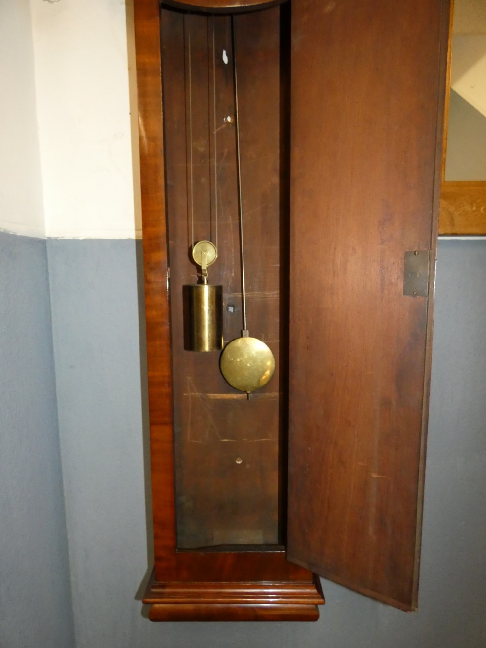 A tall Victorian mahogany regulator wall clock featuring a circular white dial with Roman numerals and a long rectangular wooden trunk. View III