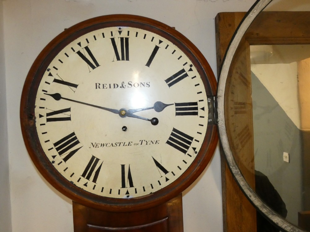 A tall Victorian mahogany regulator wall clock featuring a circular white dial with Roman numerals and a long rectangular wooden trunk. View IV