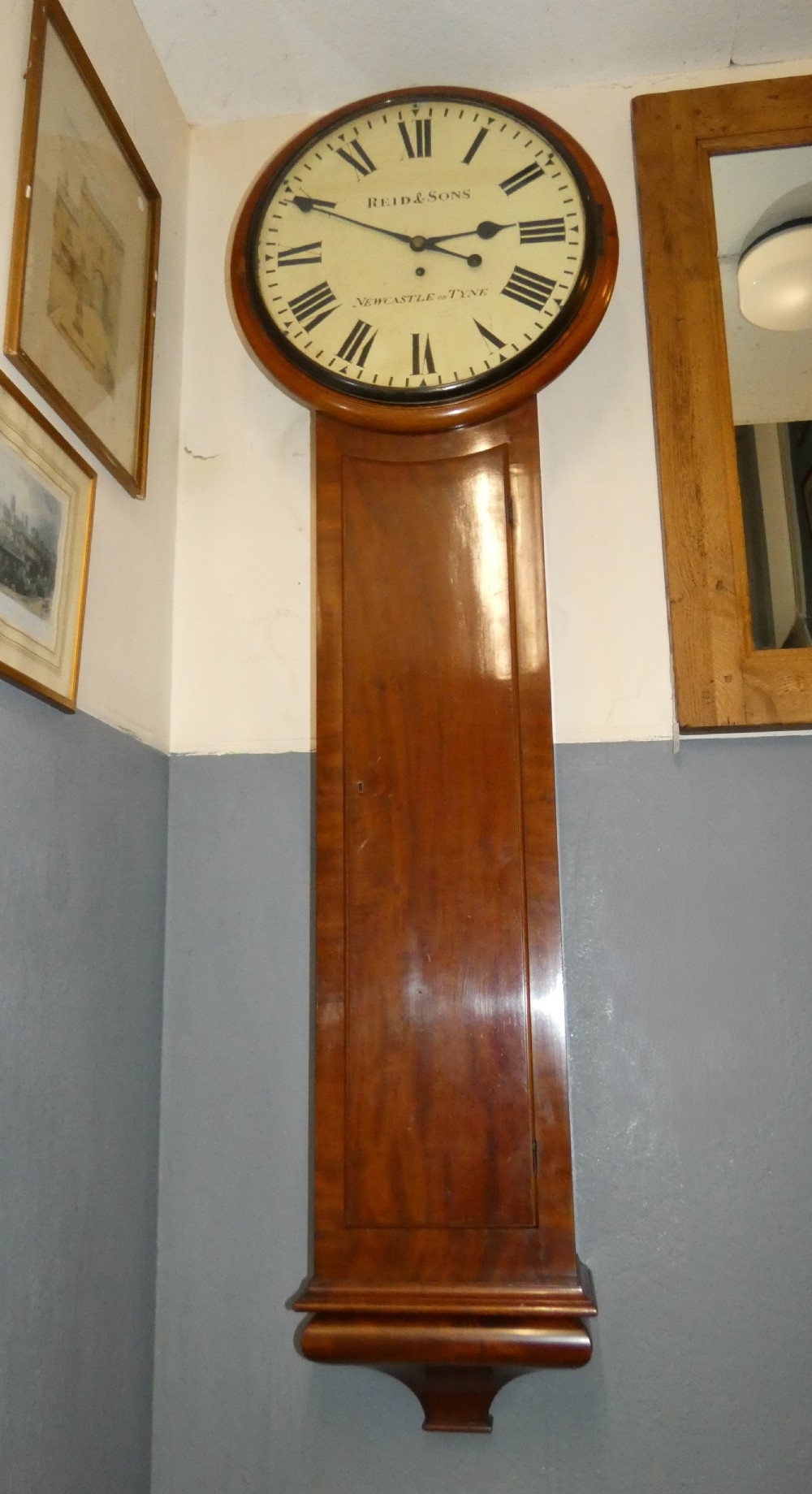 A tall Victorian mahogany regulator wall clock featuring a circular white dial with Roman numerals and a long rectangular wooden trunk. View XI