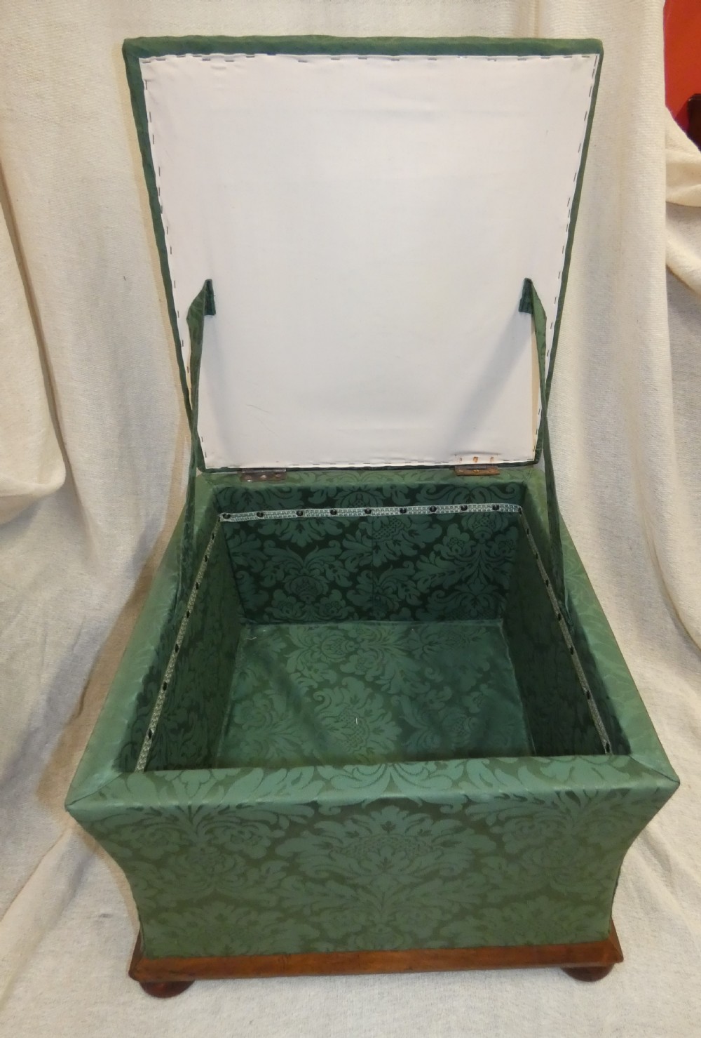 A square upholstered storage ottoman covered in green patterned damask fabric resting on a dark timber plinth base with rounded bun feet. View III