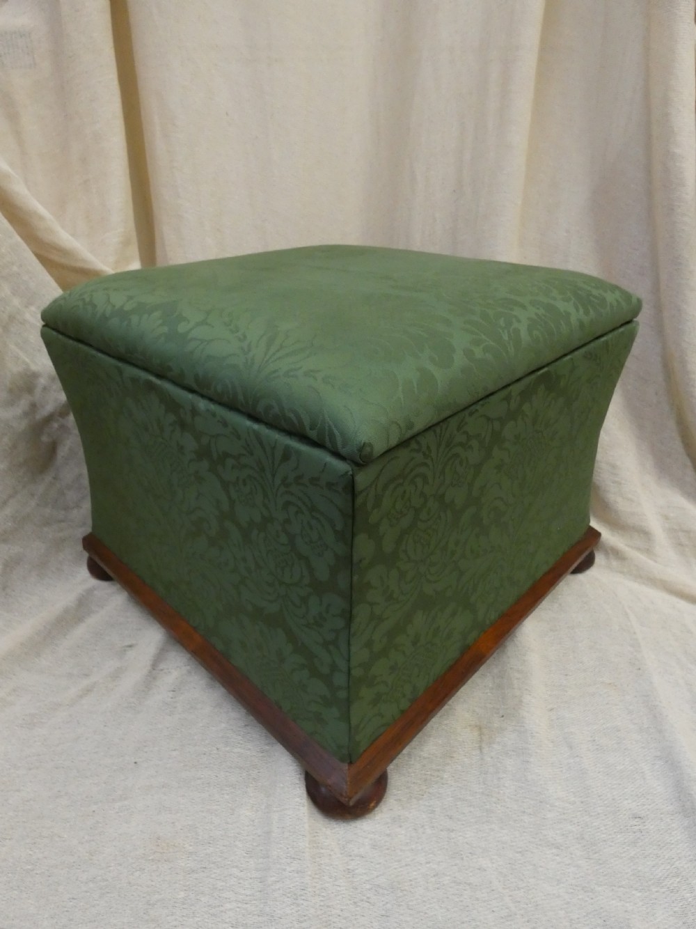 A square upholstered storage ottoman covered in green patterned damask fabric resting on a dark timber plinth base with rounded bun feet. View IV