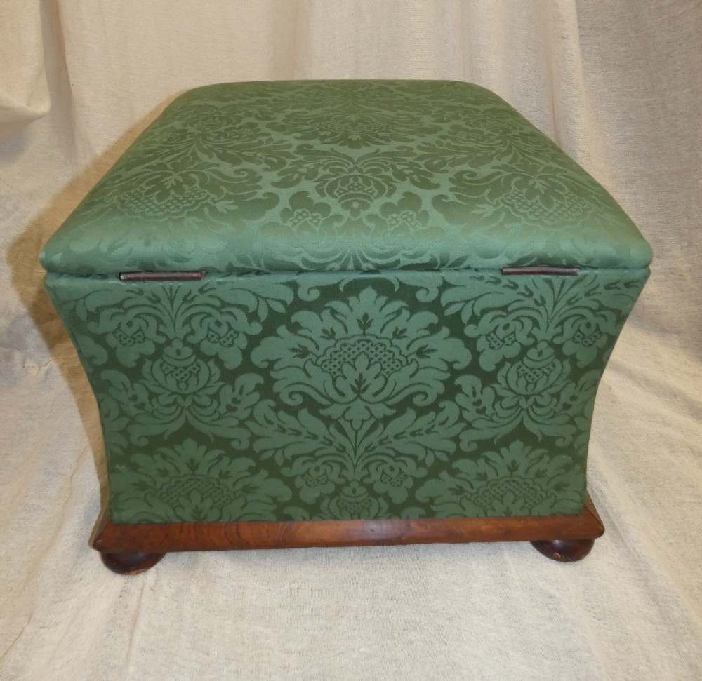 A square upholstered storage ottoman covered in green patterned damask fabric resting on a dark timber plinth base with rounded bun feet. View V
