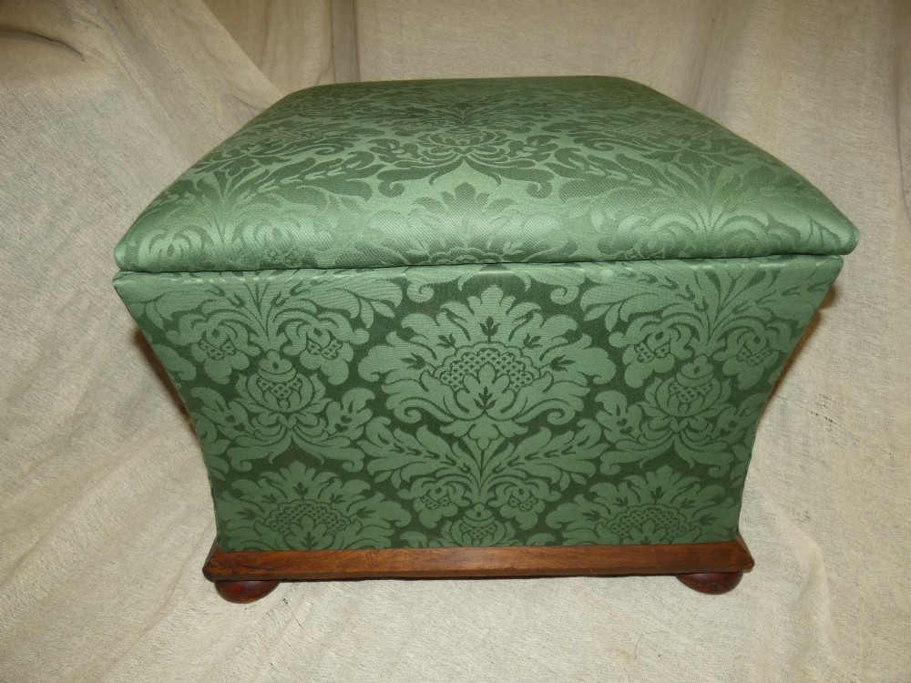 A square upholstered storage ottoman covered in green patterned damask fabric resting on a dark timber plinth base with rounded bun feet. View VII