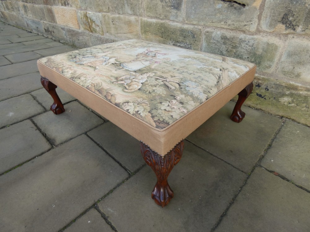 A large square upholstered centre stool featuring a tapestry top with figures in a landscape and dark carved mahogany cabriole legs with ball and claw feet.