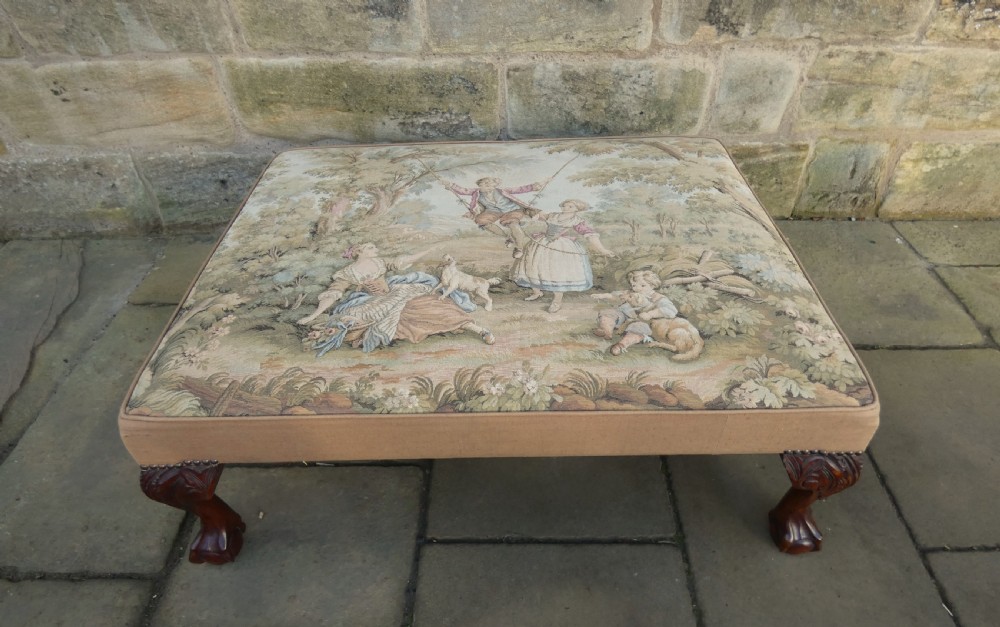 A large square upholstered centre stool featuring a tapestry top with figures in a landscape and dark carved mahogany cabriole legs with ball and claw feet. View II