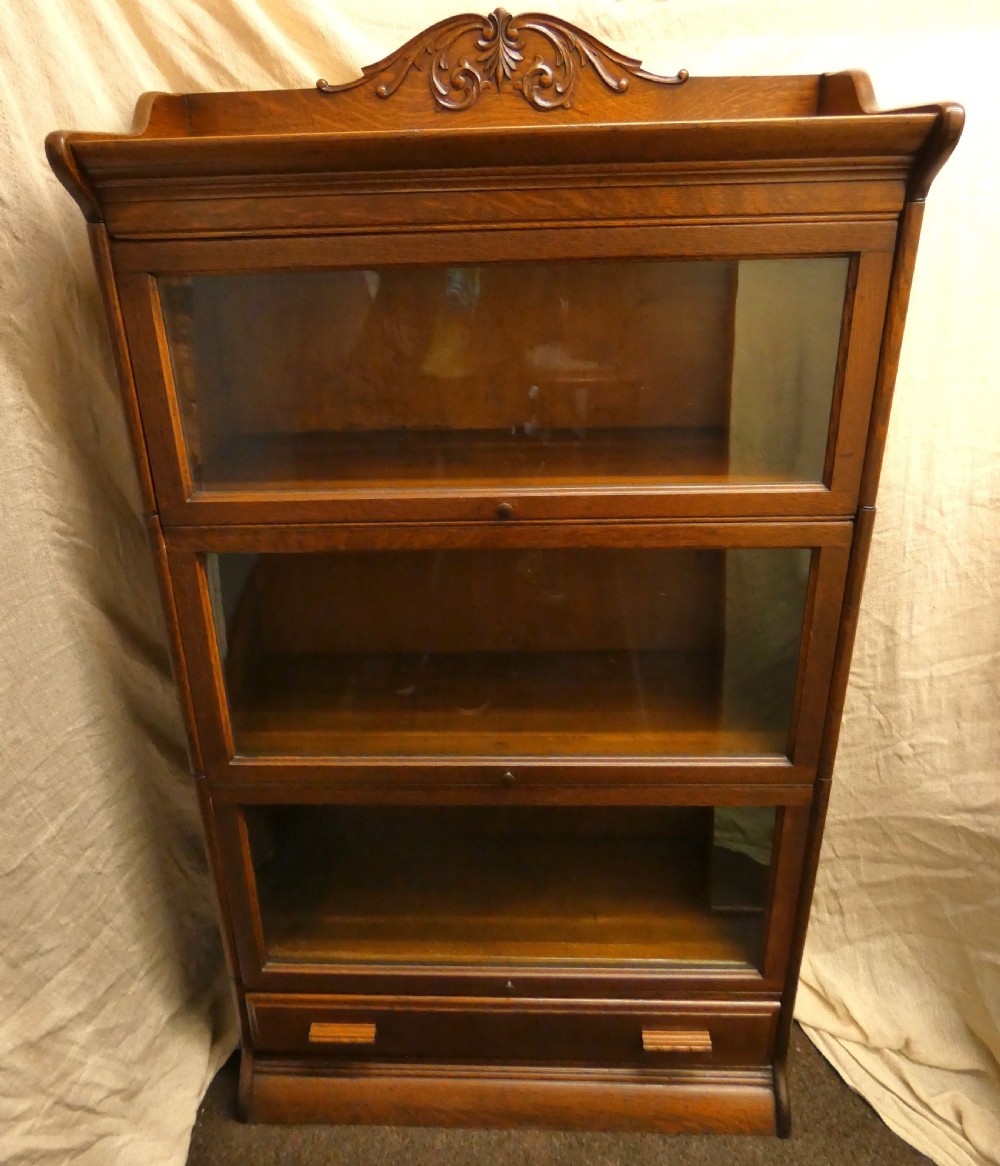 An antique medium brown oak sectional bookcase featuring three glazed lift-up doors, a carved top pediment, and a single base drawer.