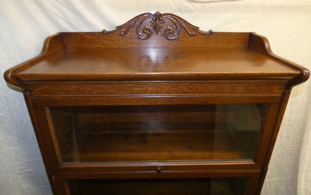 An antique medium brown oak sectional bookcase featuring three glazed lift-up doors, a carved top pediment, and a single base drawer. View II