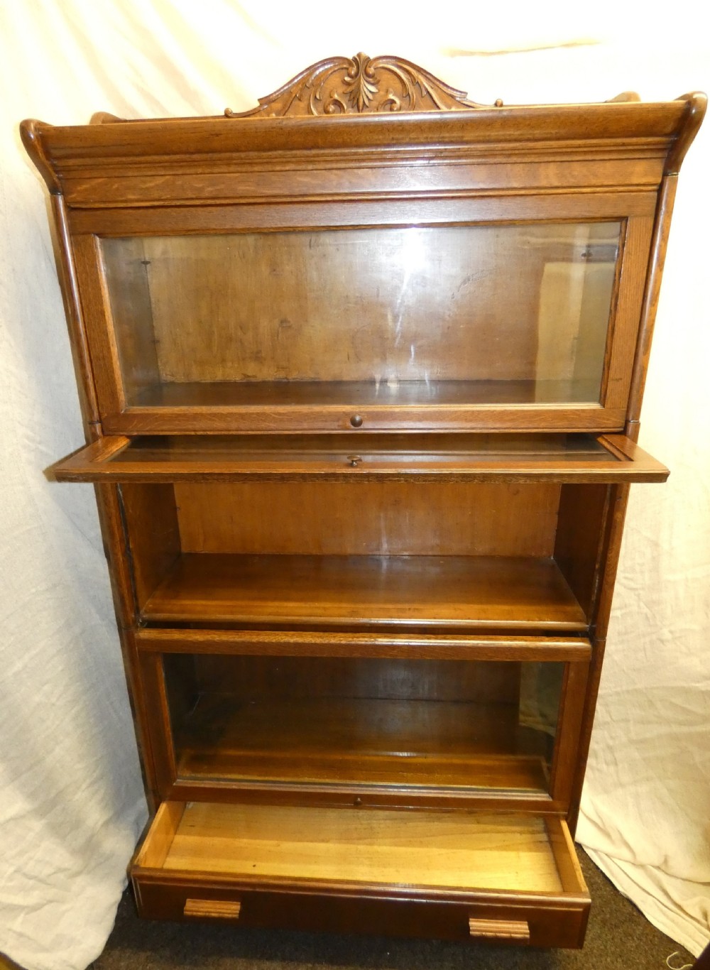 An antique medium brown oak sectional bookcase featuring three glazed lift-up doors, a carved top pediment, and a single base drawer. View III