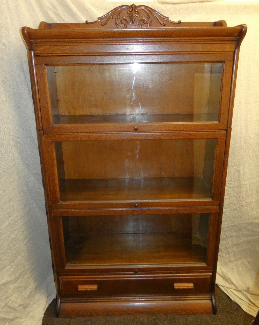 An antique medium brown oak sectional bookcase featuring three glazed lift-up doors, a carved top pediment, and a single base drawer. View IV
