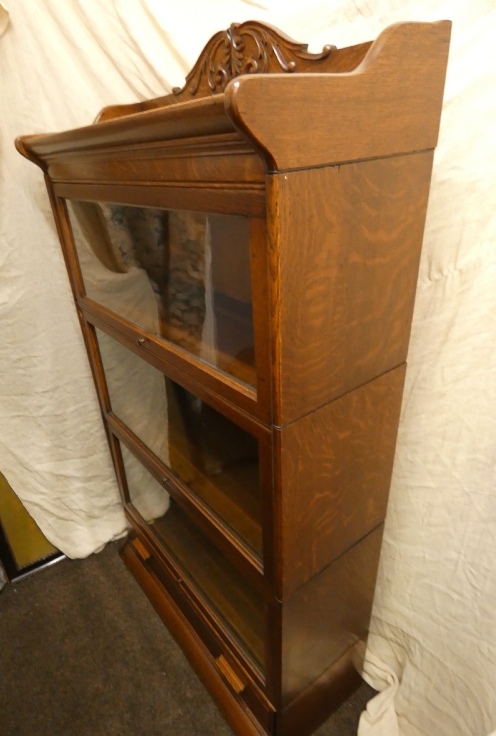 An antique medium brown oak sectional bookcase featuring three glazed lift-up doors, a carved top pediment, and a single base drawer. View V