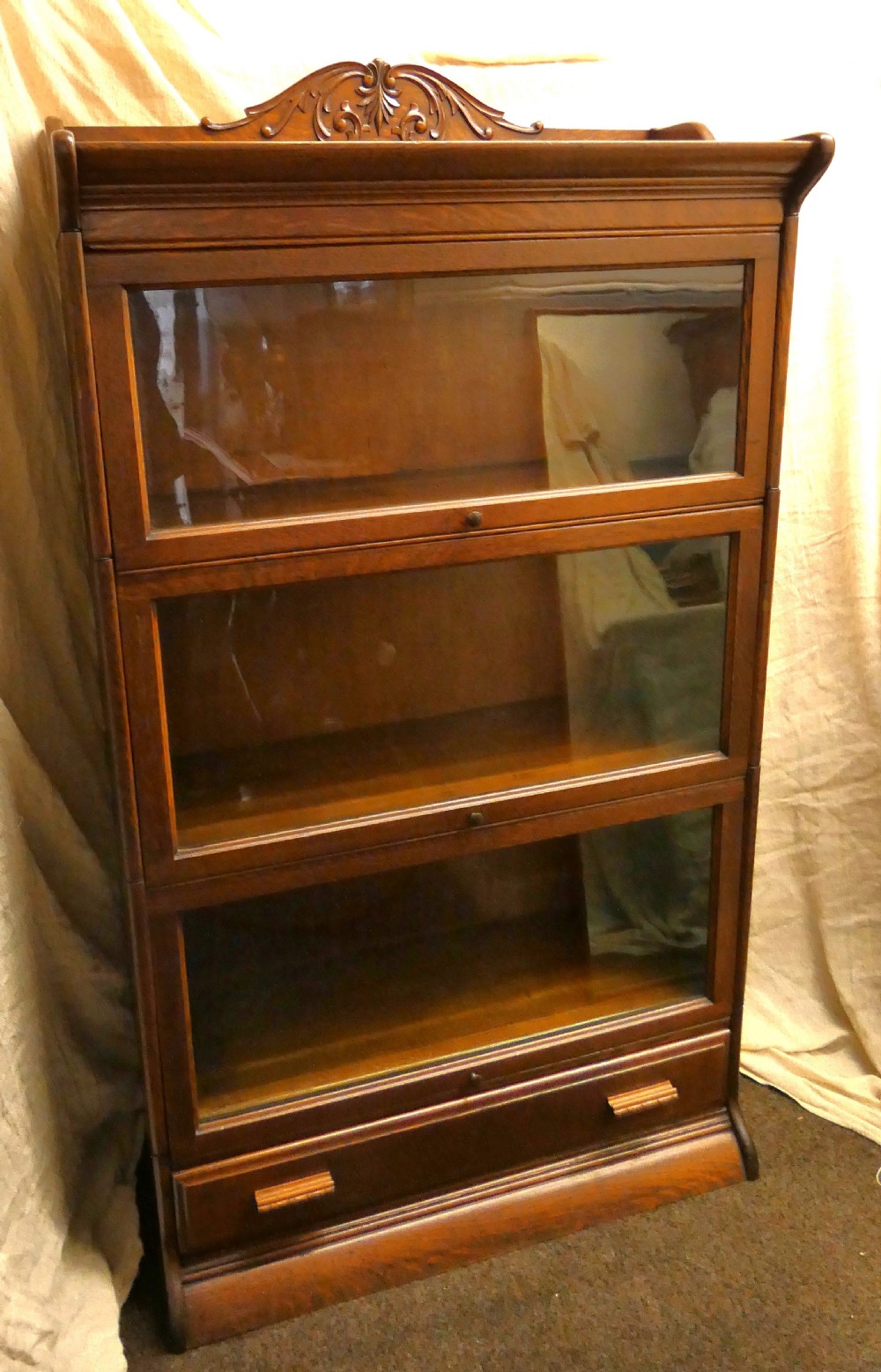 An antique medium brown oak sectional bookcase featuring three glazed lift-up doors, a carved top pediment, and a single base drawer. View VI