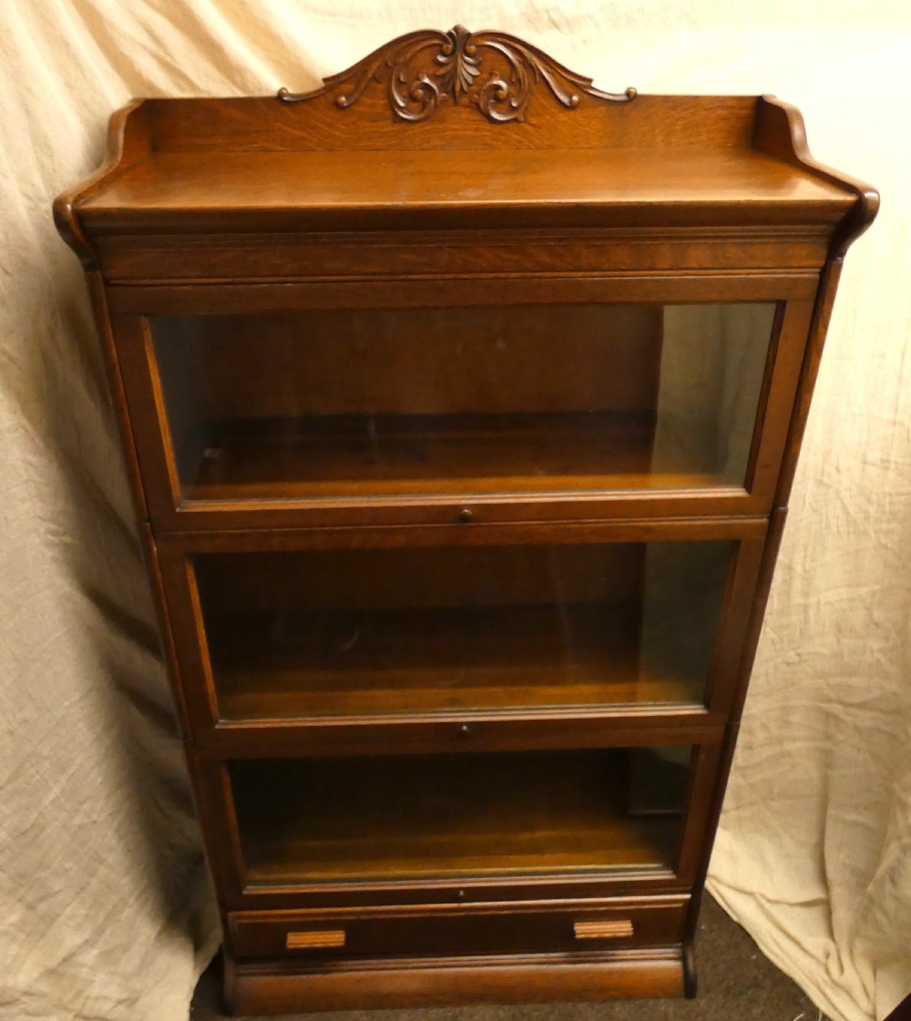 An antique medium brown oak sectional bookcase featuring three glazed lift-up doors, a carved top pediment, and a single base drawer. View VII