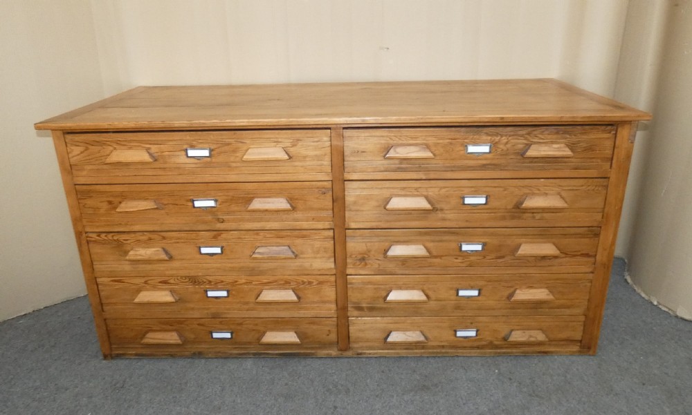 A wide pine cabinet featuring ten shallow drawers in two columns, each fitted with angular wooden handles and metal label holders.