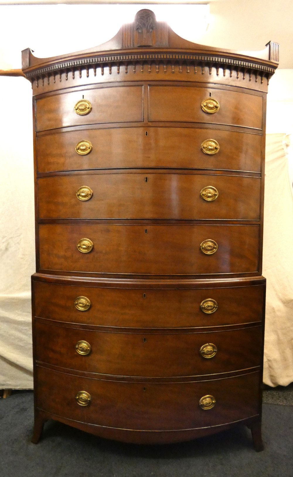 An antique bow-front mahogany chest on chest featuring graduated drawers, brass oval drop handles, and a carved pediment crown.