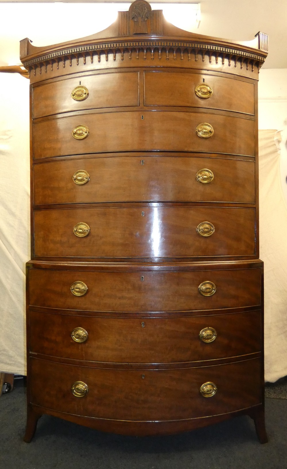 An antique bow-front mahogany chest on chest featuring graduated drawers, brass oval drop handles, and a carved pediment crown. View IV