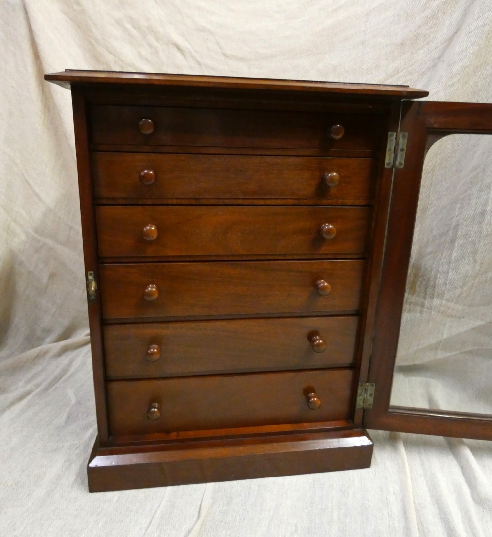 An antique warm brown mahogany specimen cabinet featuring five shallow drawers with wooden knob handles set behind a framed front on a plinth base. View III