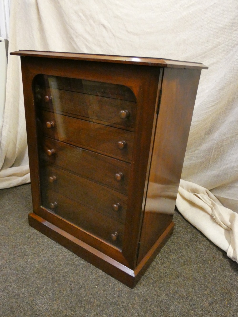An antique warm brown mahogany specimen cabinet featuring five shallow drawers with wooden knob handles set behind a framed front on a plinth base. View VI