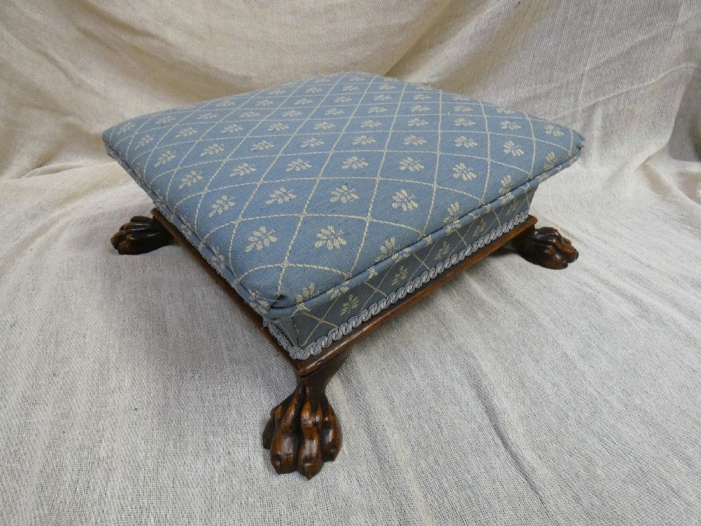 A low square upholstered footstool featuring light blue patterned fabric, decorative braid trim, and dark carved timber claw feet.