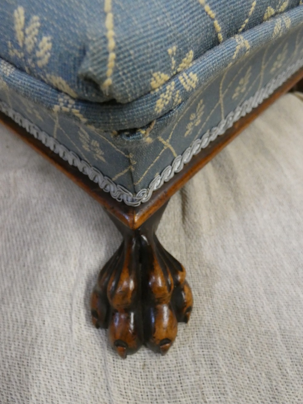 A low square upholstered footstool featuring light blue patterned fabric, decorative braid trim, and dark carved timber claw feet. View III