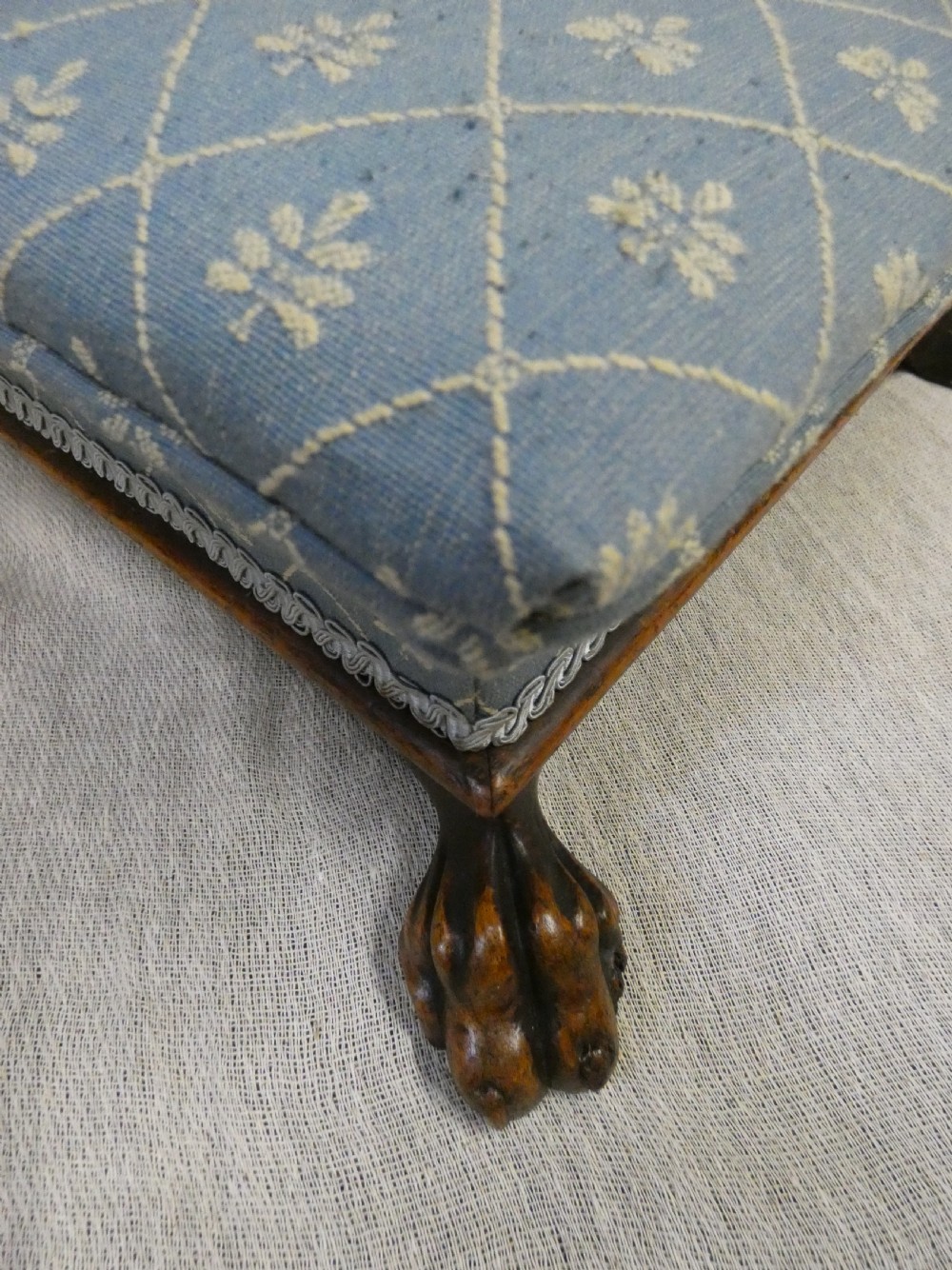 A low square upholstered footstool featuring light blue patterned fabric, decorative braid trim, and dark carved timber claw feet. View IV
