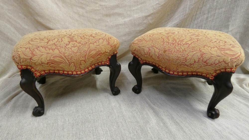 A pair of low antique footstools featuring carved dark wood cabriole legs and gold damask pattern fabric upholstery.