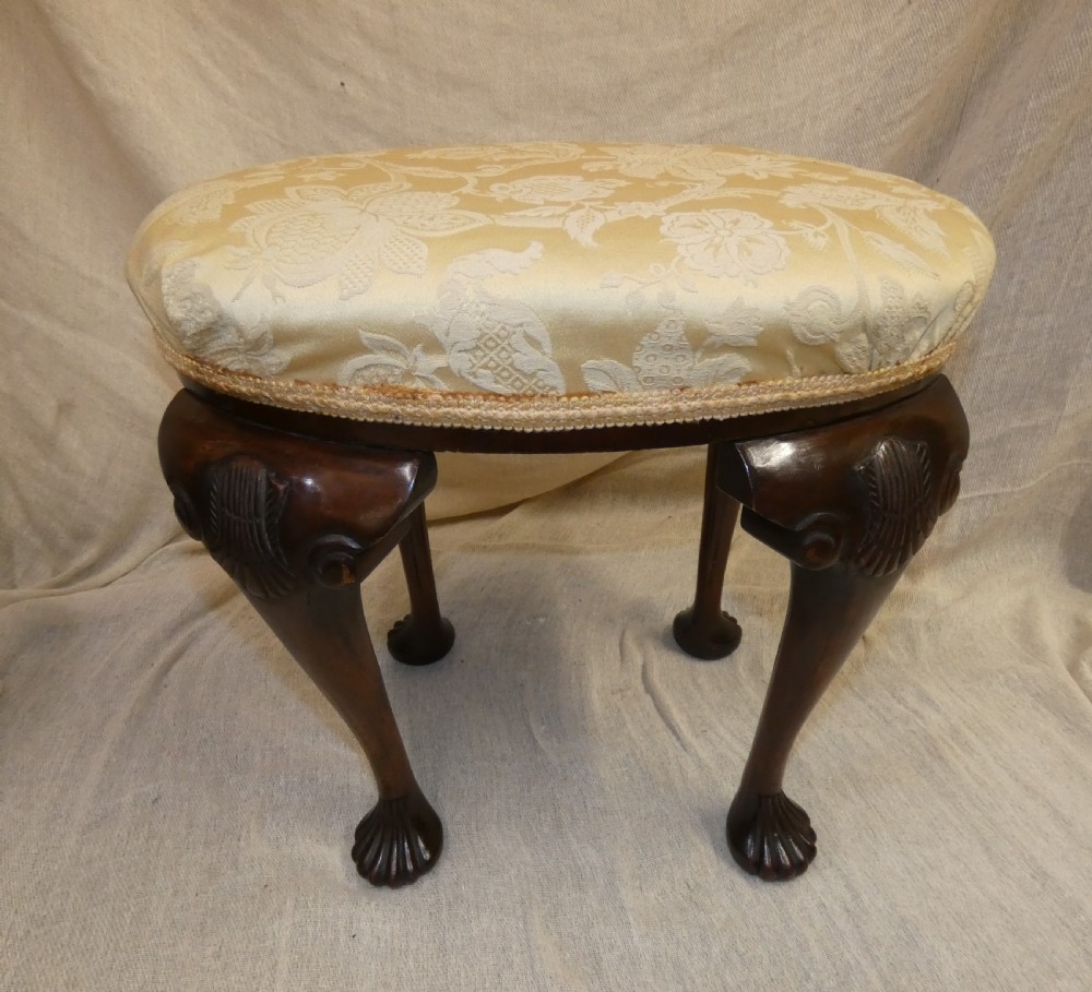 An oval upholstered antique dressing stool featuring cream damask fabric and dark mahogany cabriole legs with carved ball and claw feet.