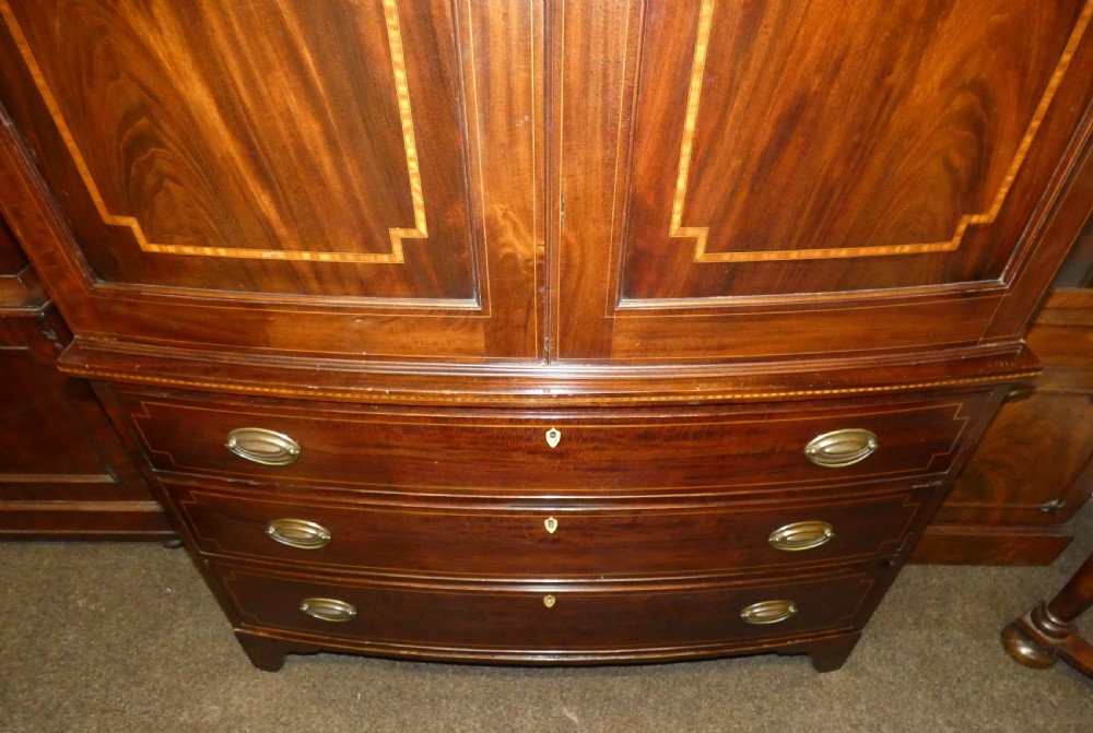 An antique mahogany bowfront linen press featuring twin panelled cupboard doors with inlay above three long drawers with brass oval handles. View V