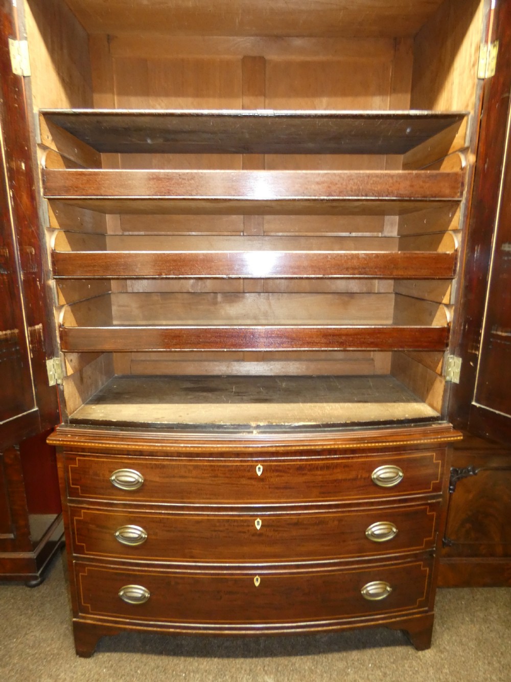 An antique mahogany bowfront linen press featuring twin panelled cupboard doors with inlay above three long drawers with brass oval handles. View VI