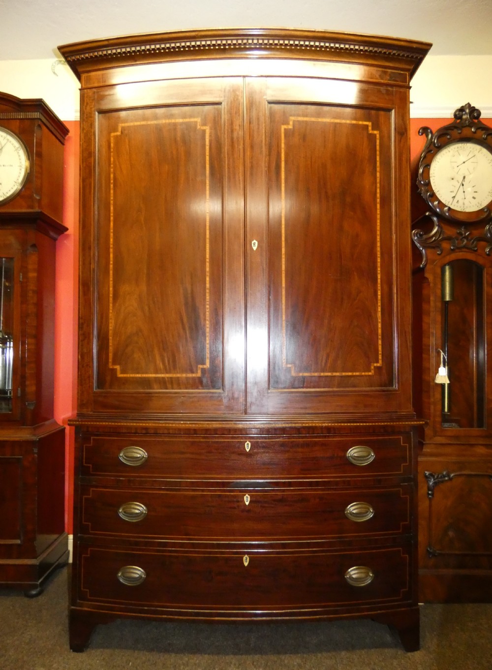 An antique mahogany bowfront linen press featuring twin panelled cupboard doors with inlay above three long drawers with brass oval handles. View VII