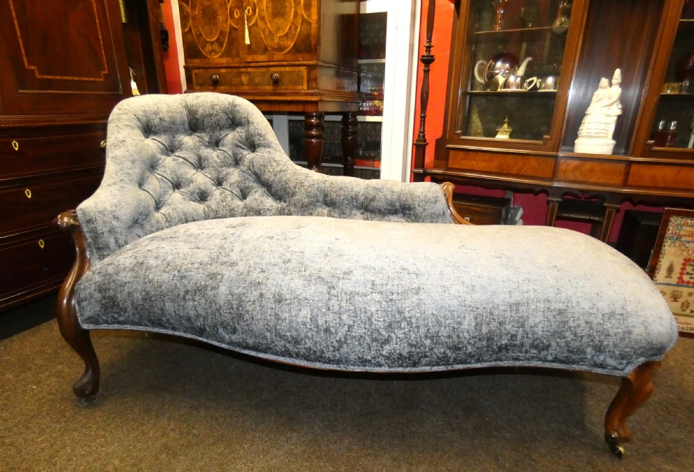 A Victorian-style chaise longue upholstered in textured grey fabric featuring a deep button-tufted asymmetrical backrest and carved wooden cabriole legs on brass castors. View II