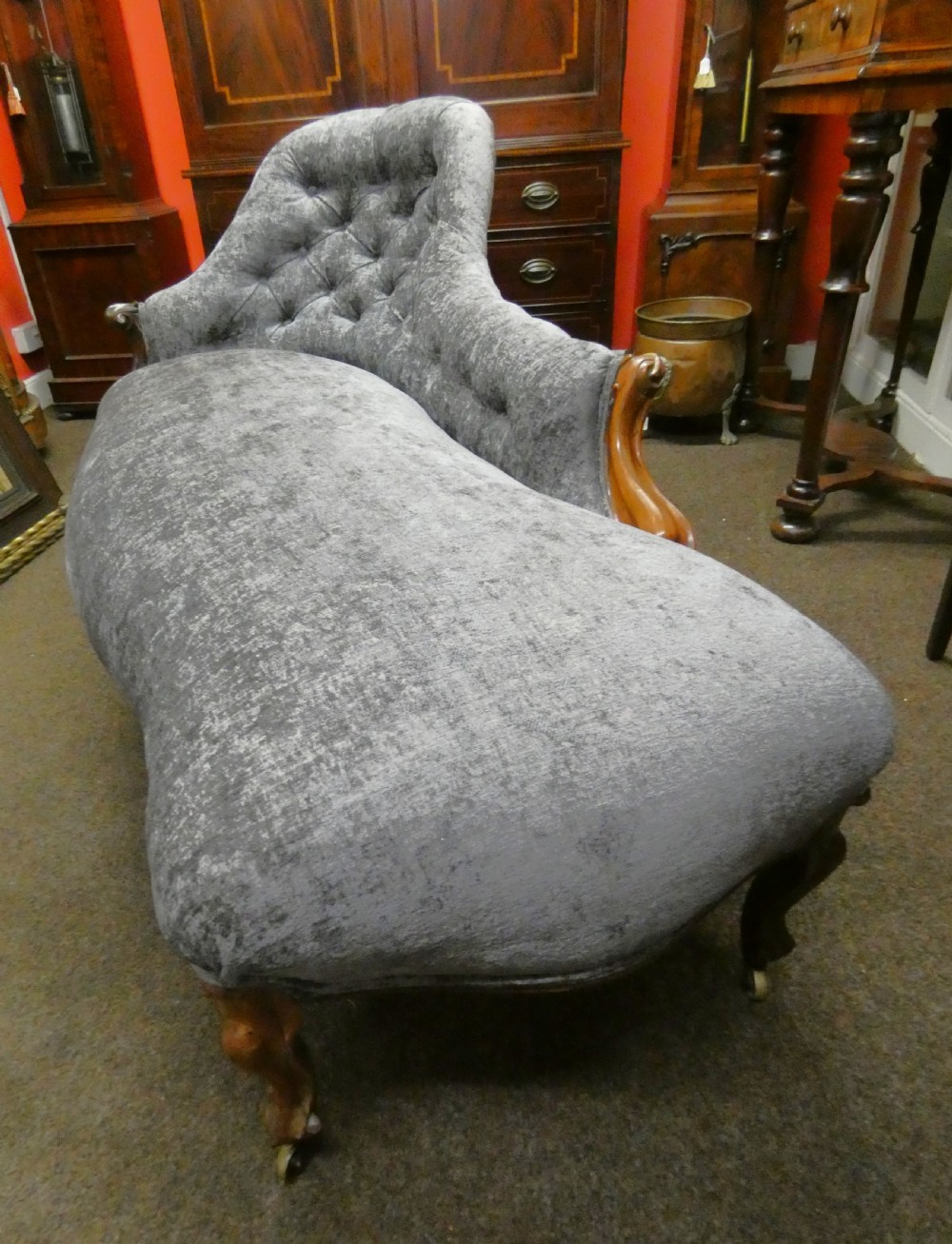 A Victorian-style chaise longue upholstered in textured grey fabric featuring a deep button-tufted asymmetrical backrest and carved wooden cabriole legs on brass castors. View VI